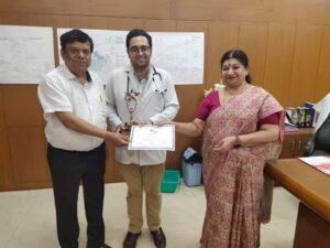 Dr. Varchasvi Mudgal receiving an award and certificate from two individuals in an office setting.