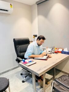 "Dr. Varchasvi Mudgal consulting with a patient in his office, writing in a notebook at his desk."