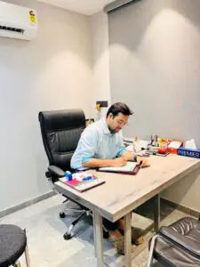 "Dr. Varchasvi Mudgal consulting with a patient in his office, writing in a notebook at his desk."